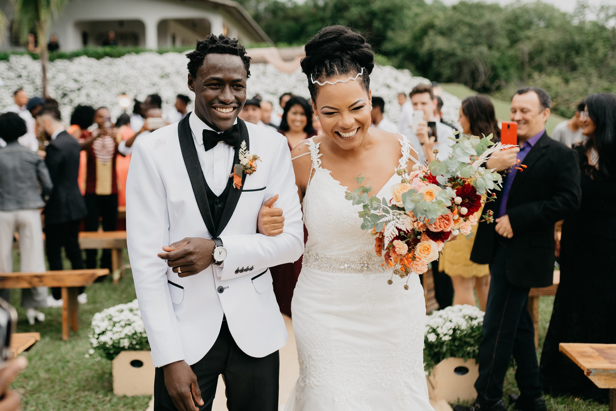 Casal sorrindo ao sair da cerimônia ao ar livre com convidados ao fundo