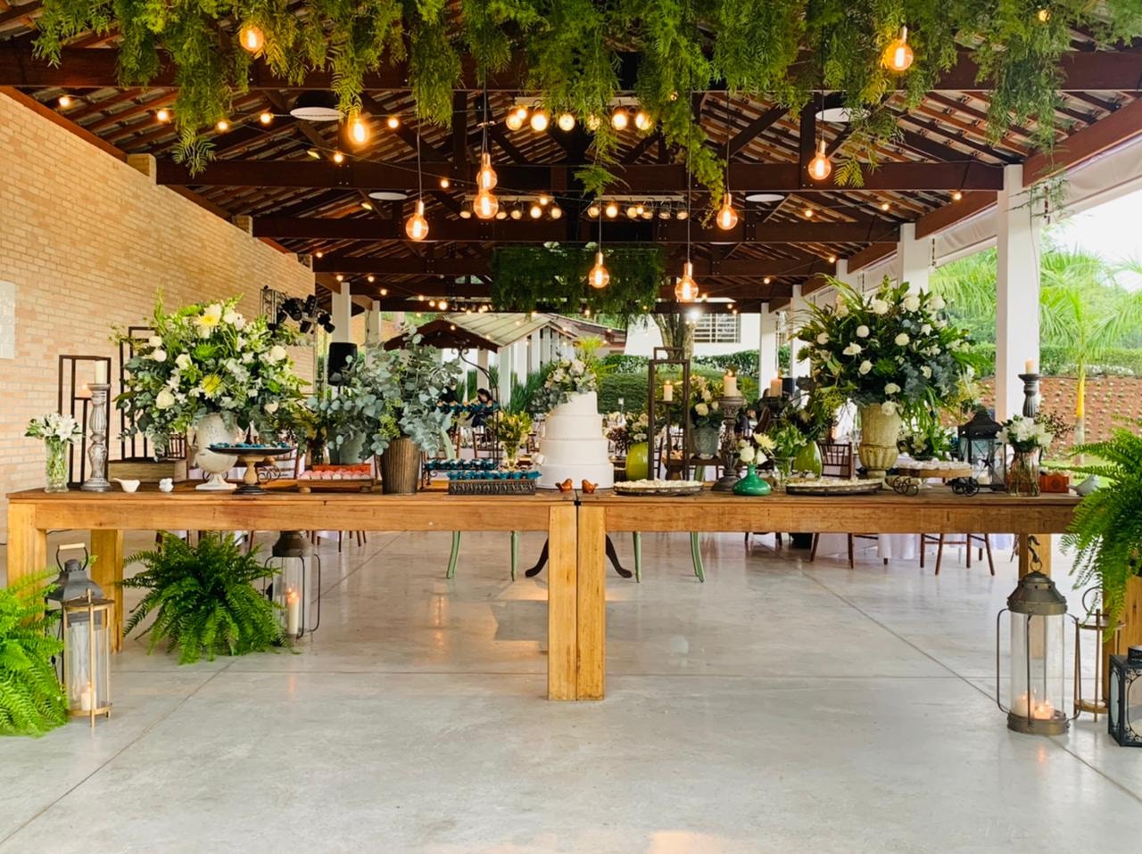 Salão decorado com vegetação suspensa, luzes pendentes e mesa de doces para casamento