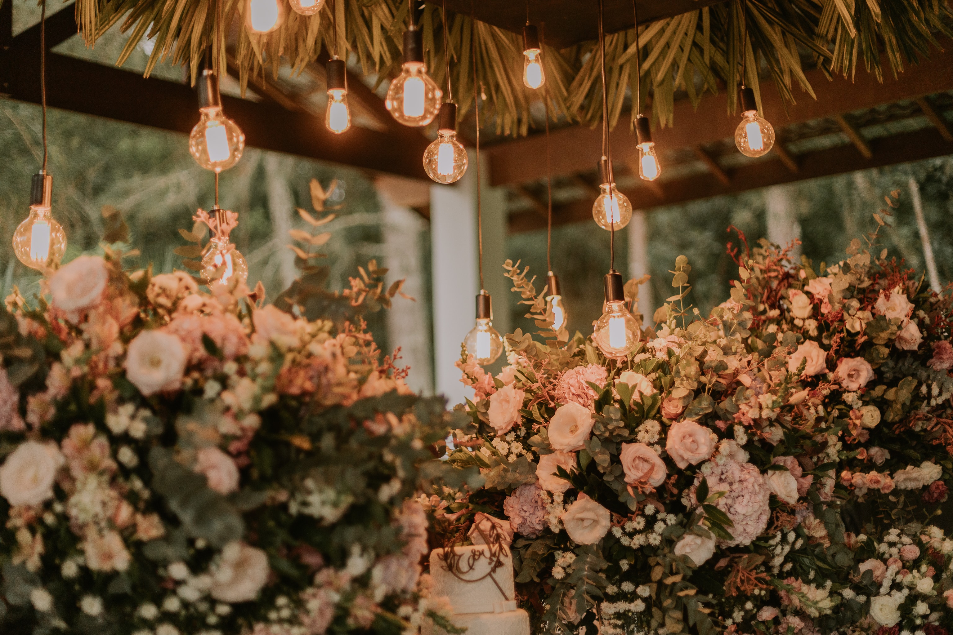 Mesa de flores rosas e brancas com luzes pendentes douradas em decoração de casamento