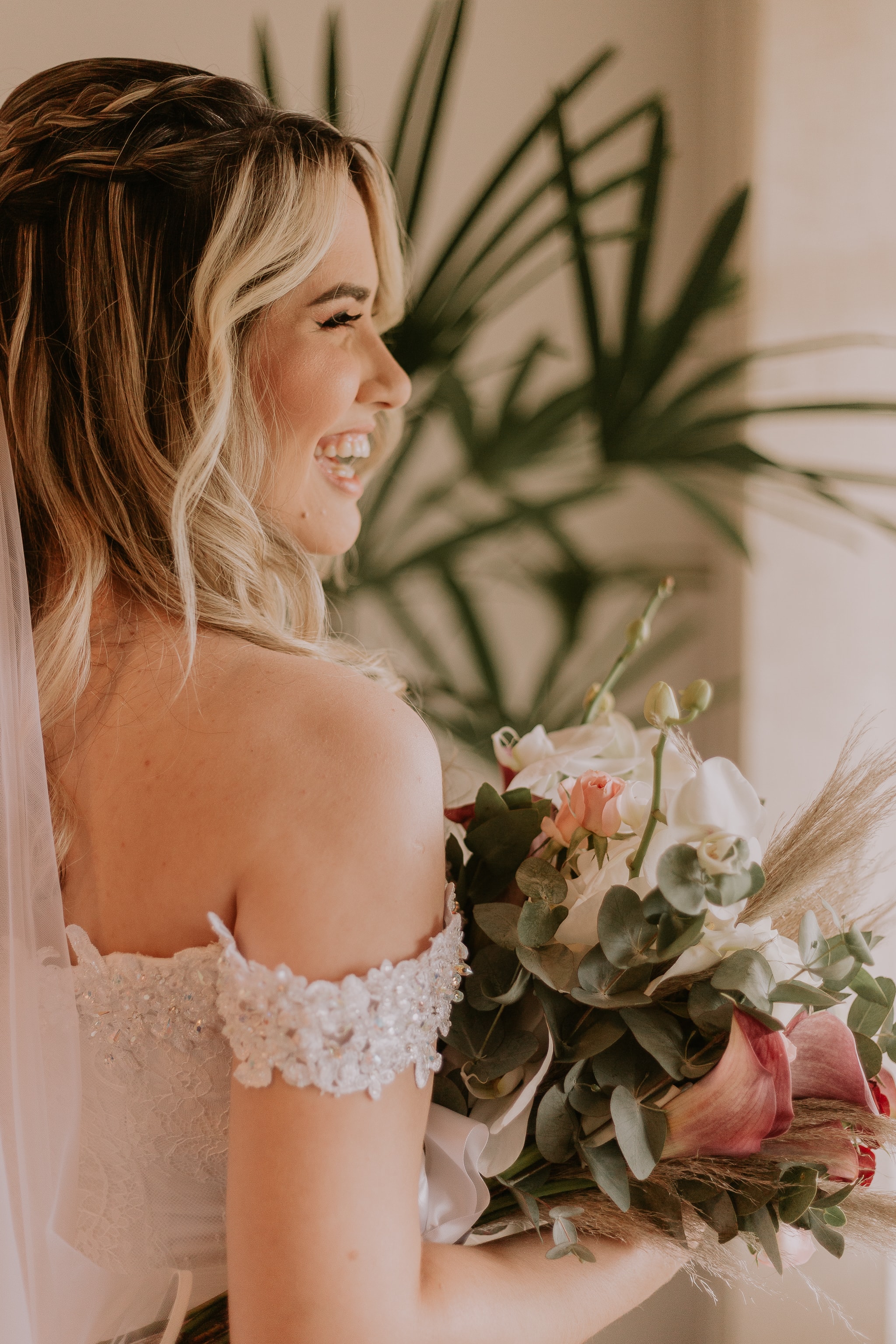 Noiva sorrindo de perfil com buquê de orquídeas e rosas em vestido off-shoulder