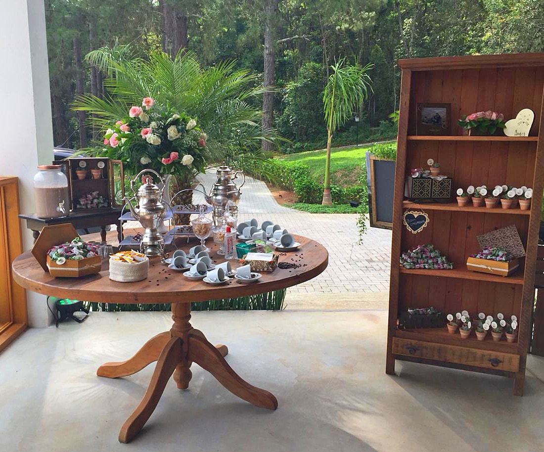 Mesa redonda de madeira com café e doces ao lado de estante com lembranças de casamento
