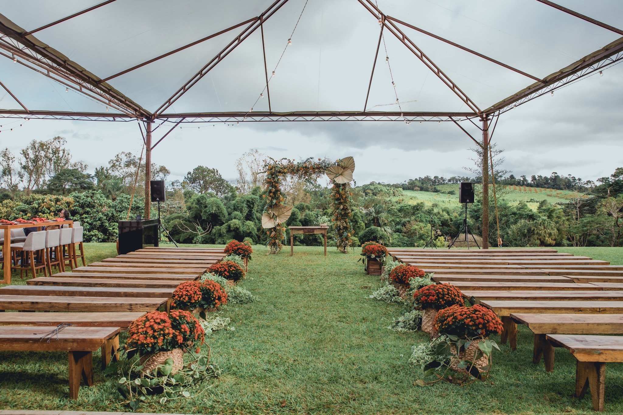 Cerimônia ao ar livre sob tenda metálica com bancos de madeira, flores laranja e arco floral com vista para as colinas