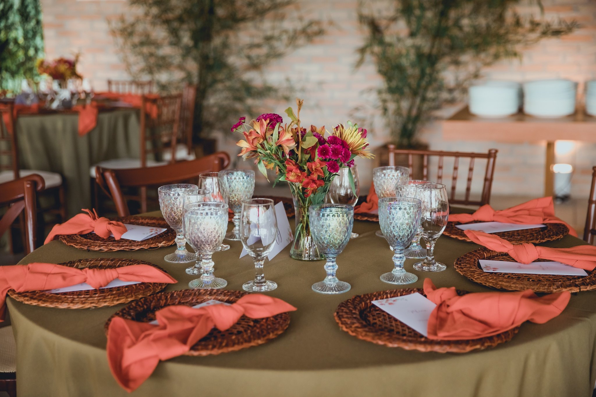 Mesa redonda decorada com guardanapos coral, taças de cristal e arranjo de flores coloridas em salão rústico