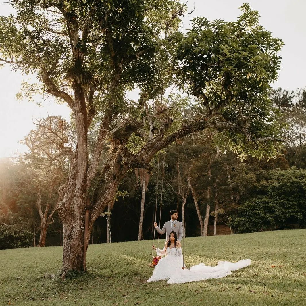 Noiva sentada em balanço sob árvore centenária com noivo ao lado ao pôr do sol no gramado do Eco Espaço