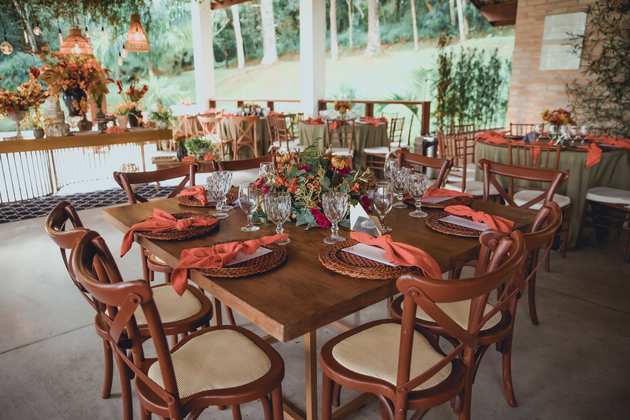 Mesa quadrada de madeira com cadeiras cross-back, guardanapos terracota e arranjo floral tropical no salão aberto