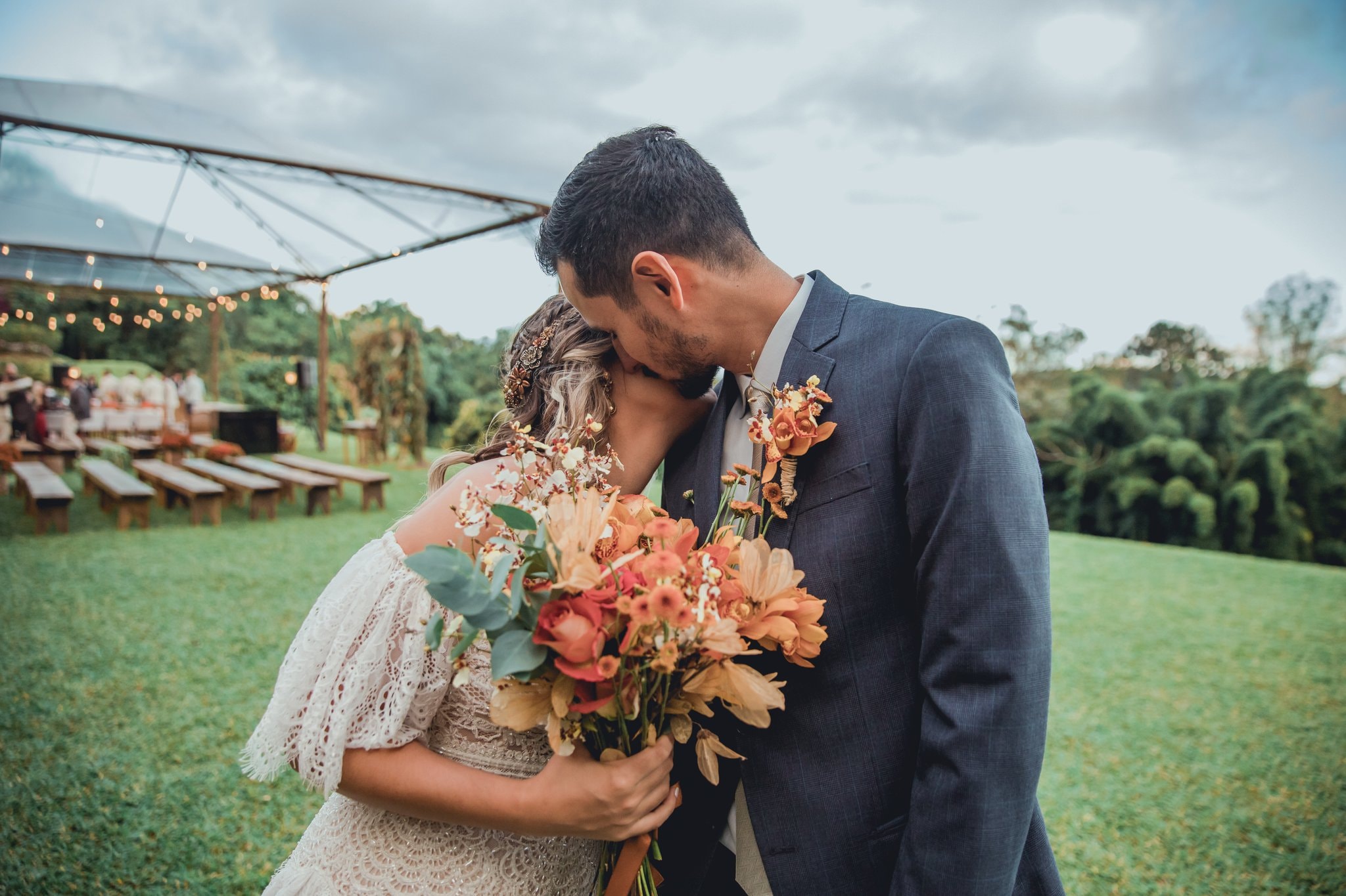 Casal de noivos se beijando no gramado sob tenda com luzes de festa e bancos de madeira ao fundo