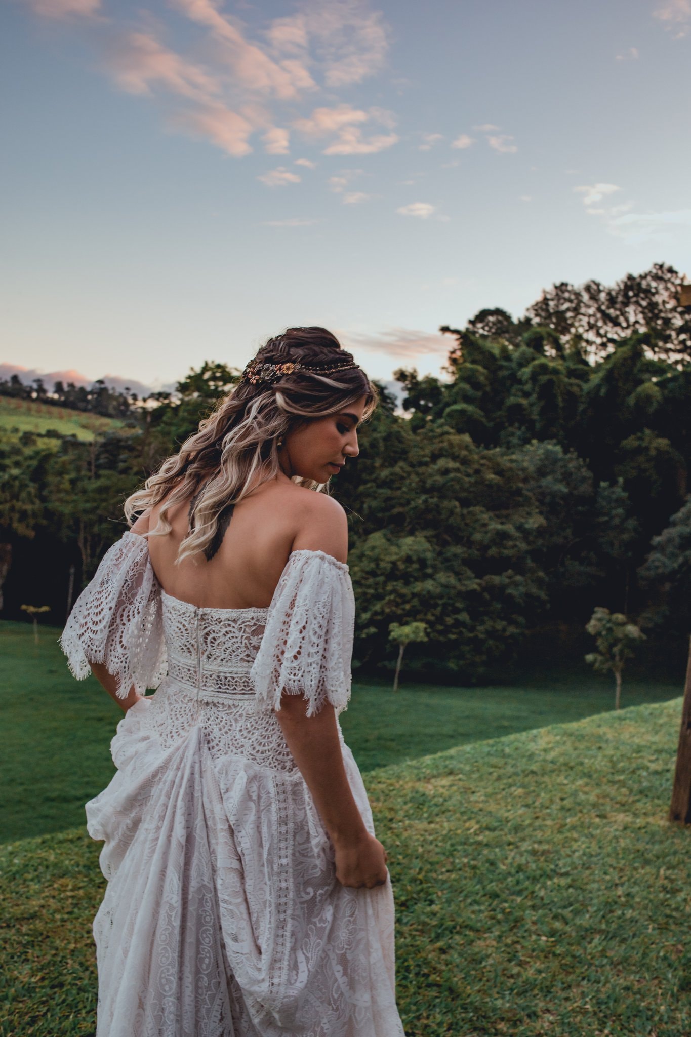 Noiva de costas com vestido de renda off-shoulder e tiara floral no gramado do Eco Espaço ao pôr do sol