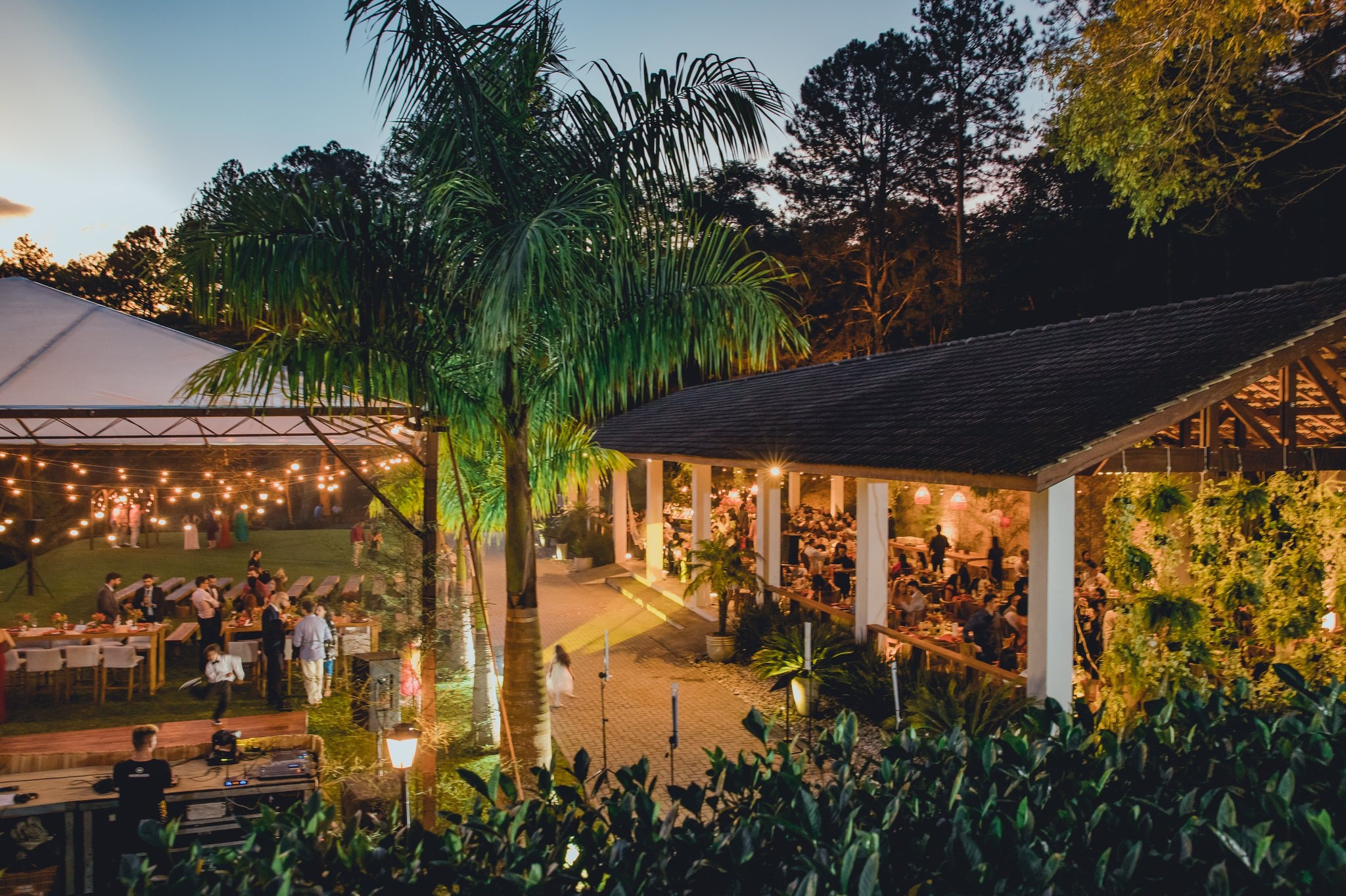 Vista panorâmica noturna do Eco Espaço com tenda, salão coberto, palmeira imperial, convidados e jardins iluminados